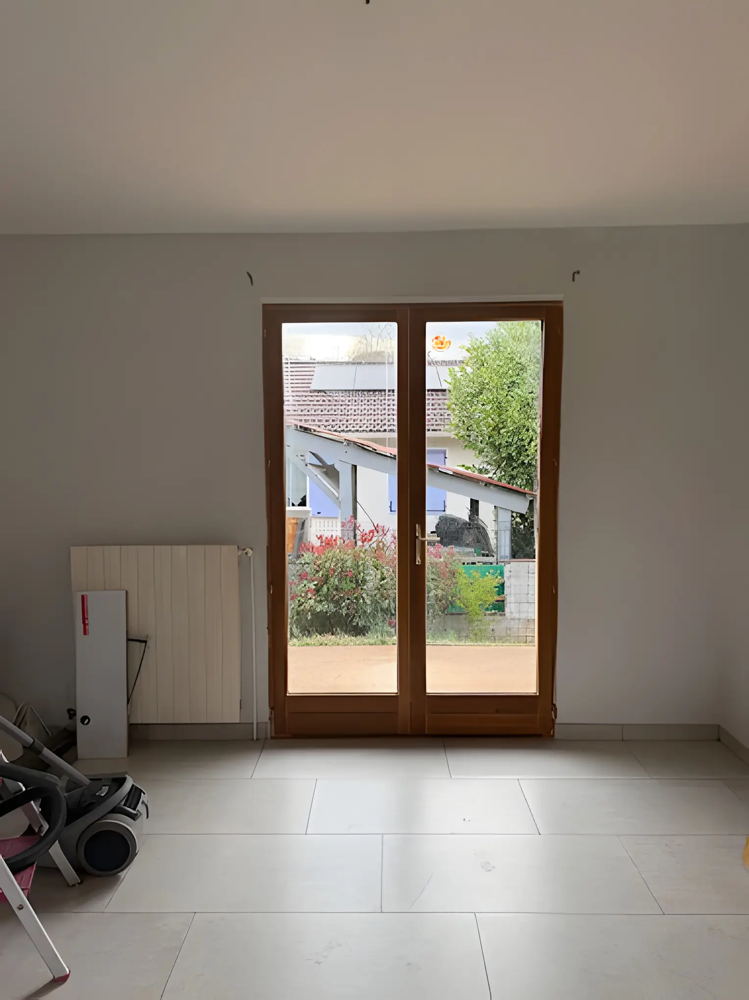 Porte-fenêtre ton bois au fond d’une pièce carrelée en travaux, jardin derrière